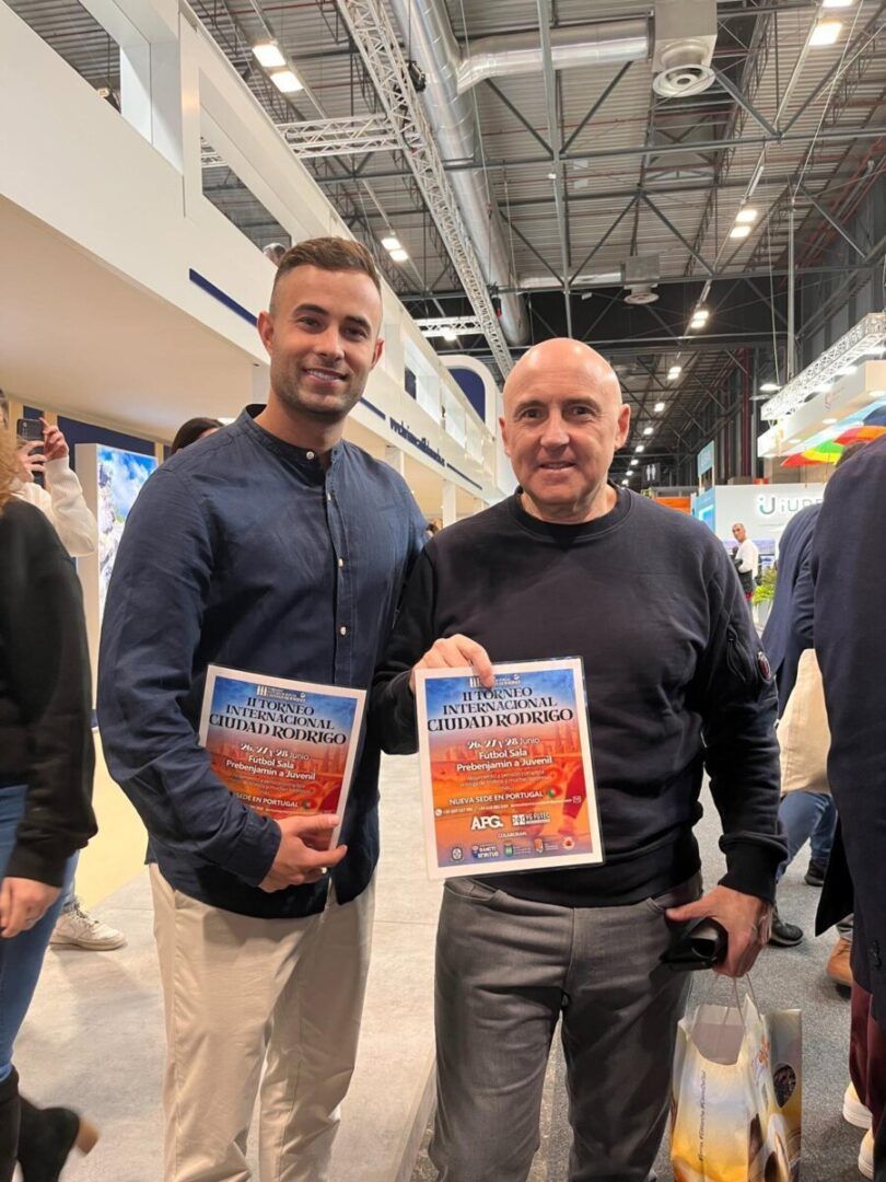 Dos hombres sosteniendo carteles del Torneo Internacional Ciudad Rodrigo 2026 en FITUR
