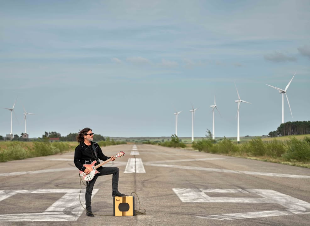 Quique González tocando la guitarra en un paisaje abierto con molinos de viento