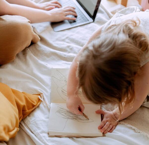 Niña dibujando en un cuaderno mientras un adulto usa una computadora portátil.