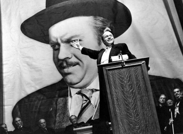 Orson Welles hablando en un mitin con un gran retrato detrás