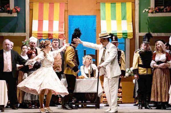 Escena de la ópera L'Elisir D'amore con varios cantantes en el escenario.
