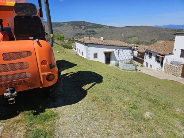 Máquina desbrozadora en un camino rural con casas blancas al fondo