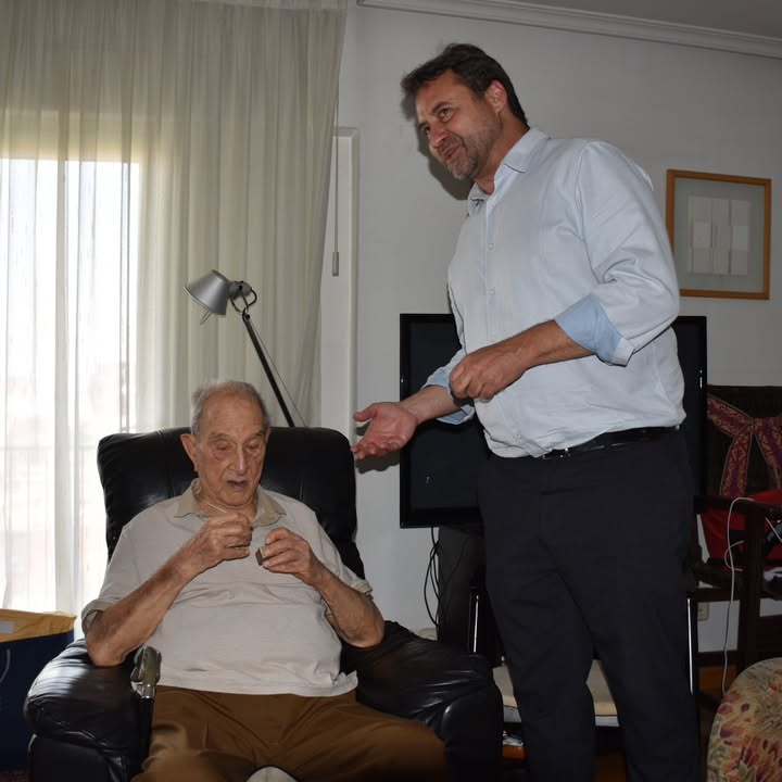 Dos hombres conversando sobre una capa histórica en un entorno doméstico.