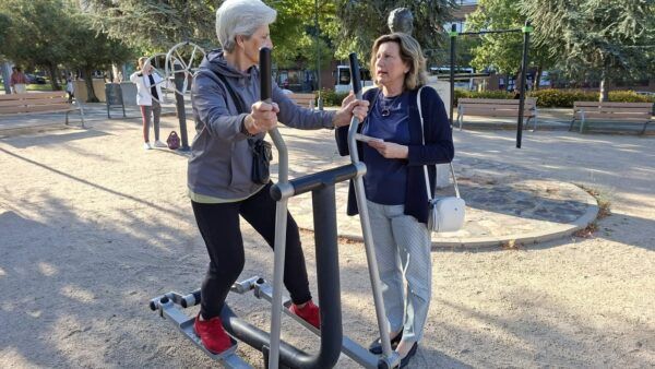 Mujer usando máquina de ejercicio en parque biosaludable con otra mujer conversando.