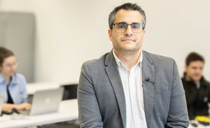 Profesor Javier Parra sentado en un aula con estudiantes al fondo
