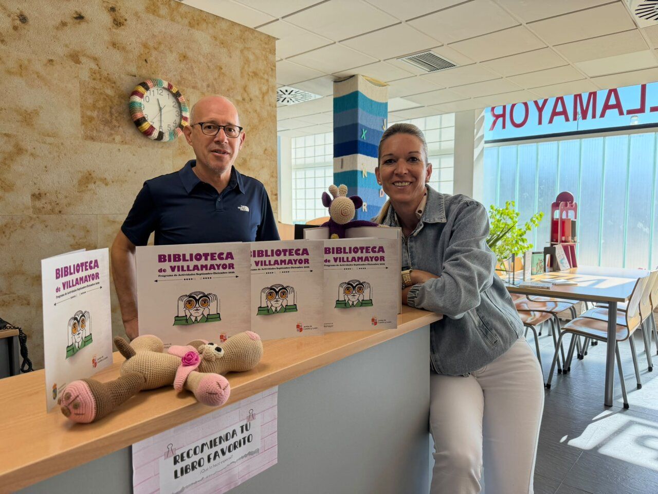 Dos personas en la biblioteca de Villamayor con libros y un muñeco de peluche