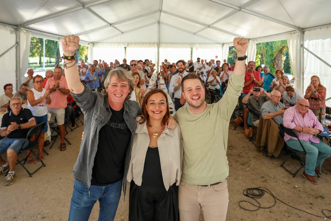 Tres personas levantando los brazos en un evento político con público al fondo.