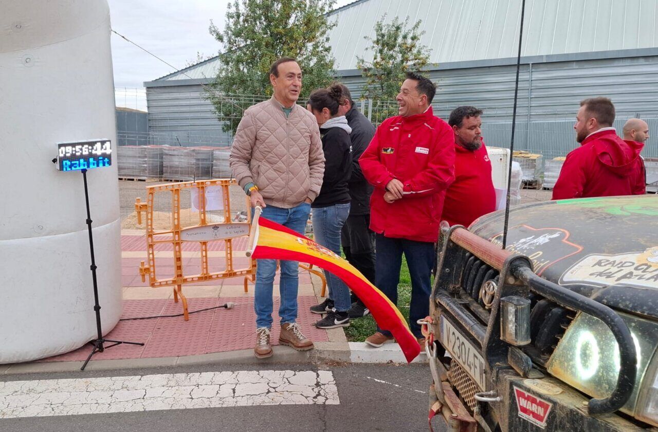 Un hombre sostiene una bandera española junto a un vehículo clásico en una línea de salida.