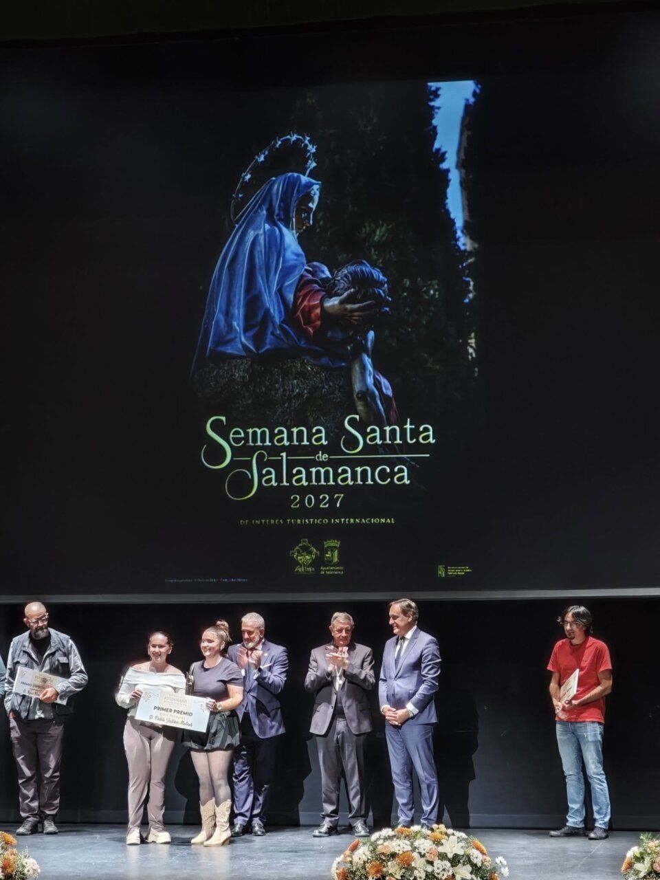 Personas en un escenario bajo el cartel de Semana Santa de Salamanca 2027.