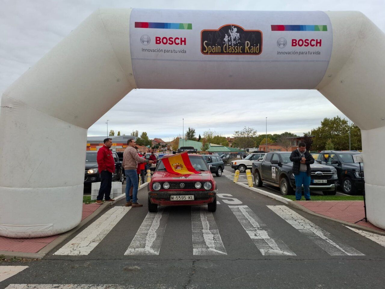 Un coche clásico rojo bajo el arco del Spain Classic Raid, rodeado de otros vehículos.