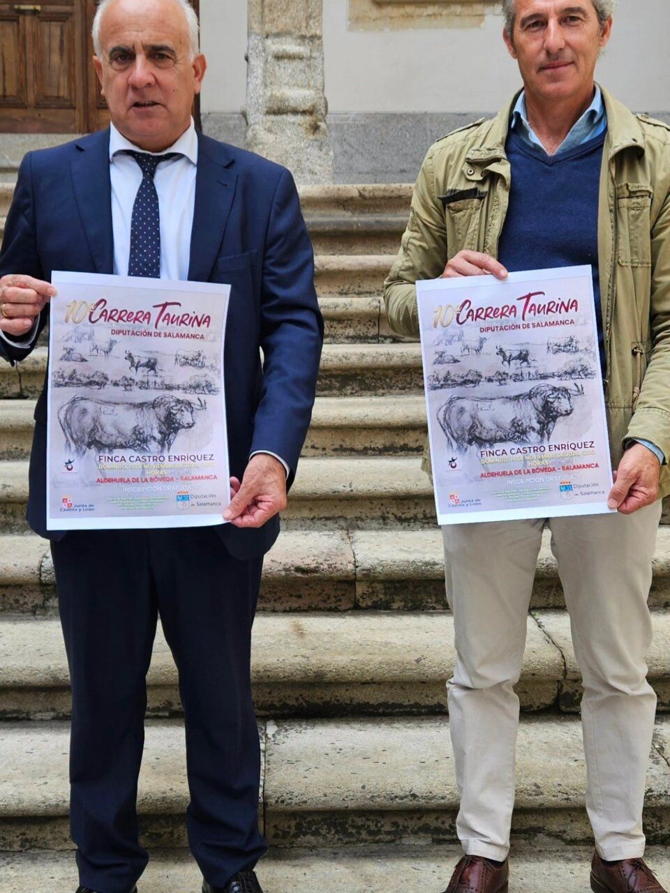 Dos hombres sostienen carteles de la X Carrera Taurina frente a una escalinata.