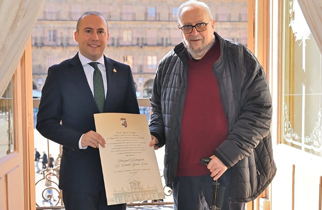 Dos hombres sostienen un diploma frente a una ventana con edificios al fondo.