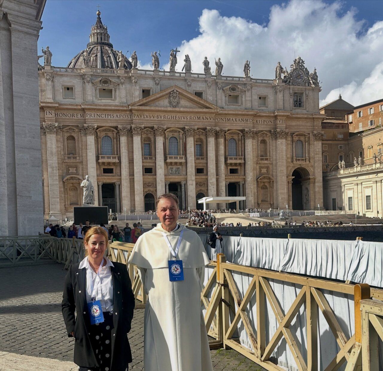 Dos personas posan ante la basílica de San Pedro bajo un cielo parcialmente nublado.