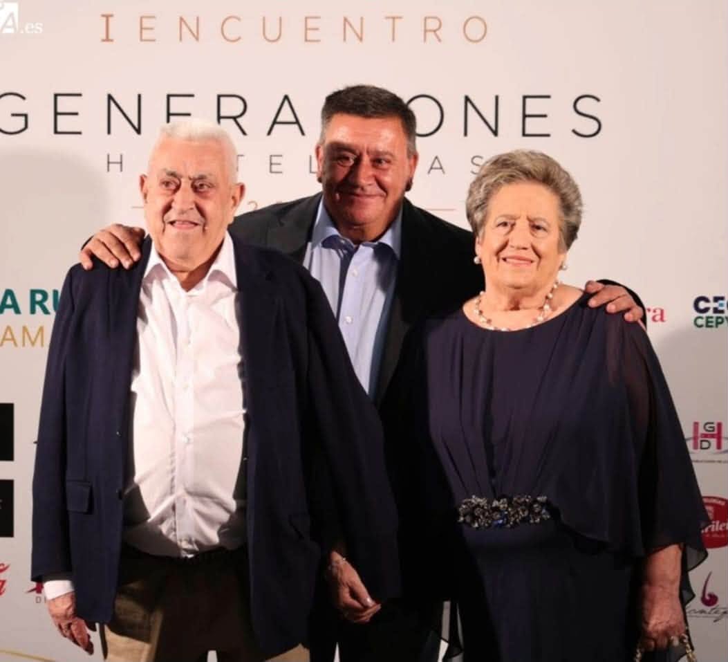 Tres personas posan juntas frente a un panel del I Encuentro de Generaciones Hosteleras.