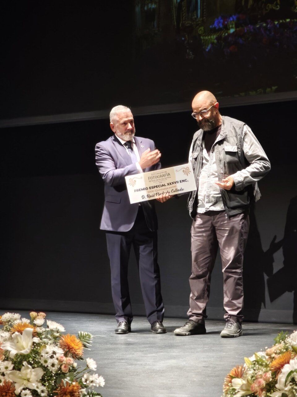 Dos hombres sostienen un diploma sobre el escenario, junto a arreglos florales.