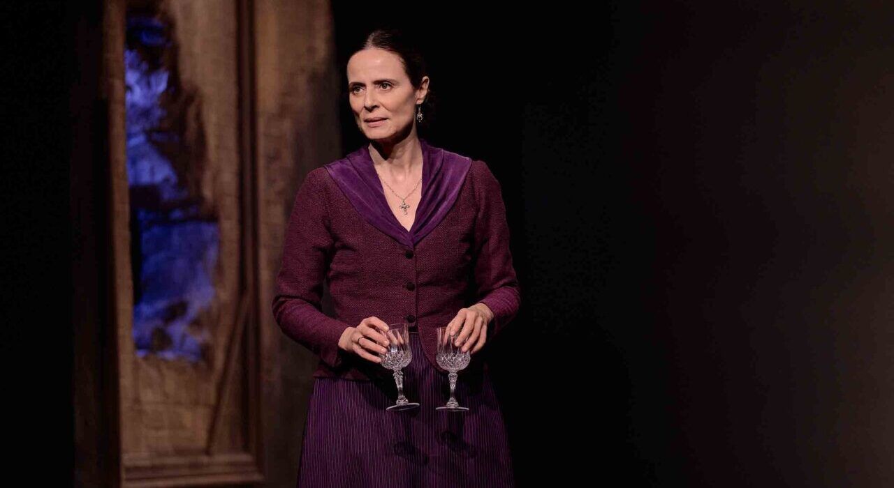 Mujer vestida de morado sostiene dos copas en una escena teatral.