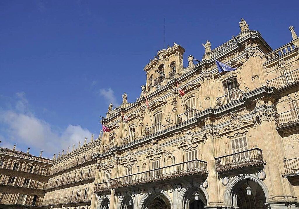 Fachada monumental de piedra con balcones, esculturas y banderas bajo un cielo azul