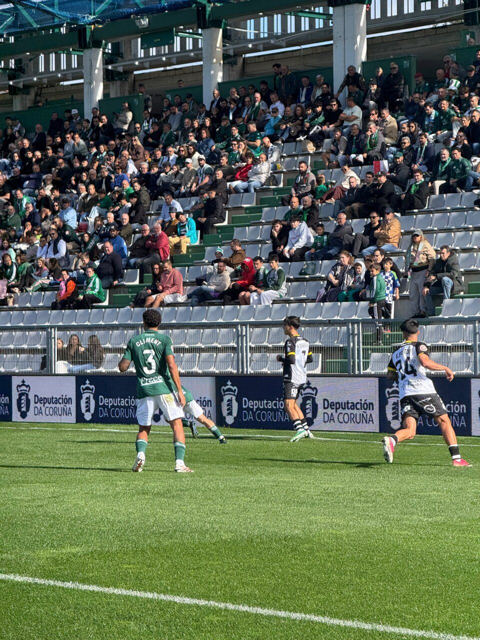 Futbolistas de verde y blanco y negro disputan el balón frente a las gradas.