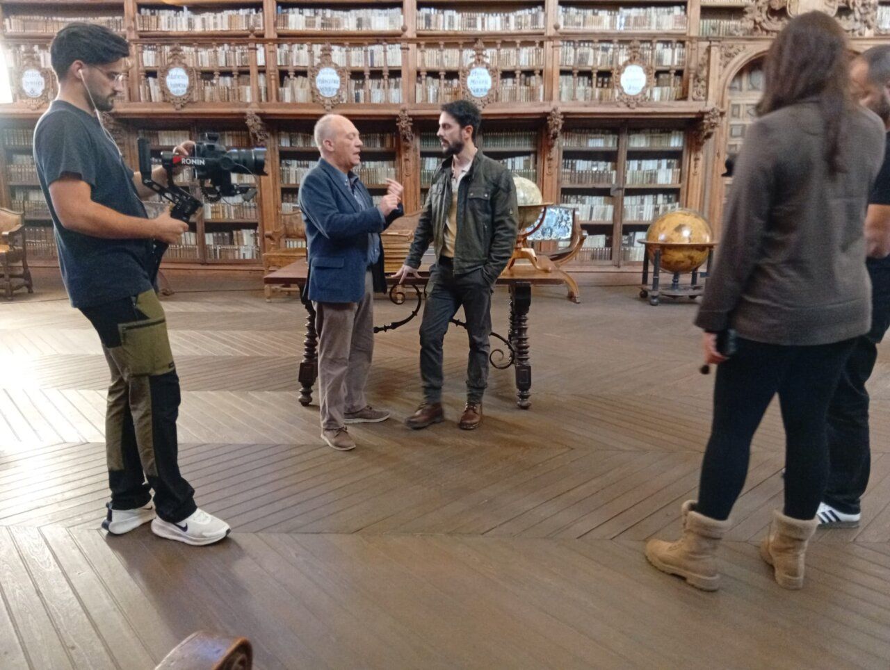 Un cámara graba a dos hombres conversando en una sala con estanterías de libros.