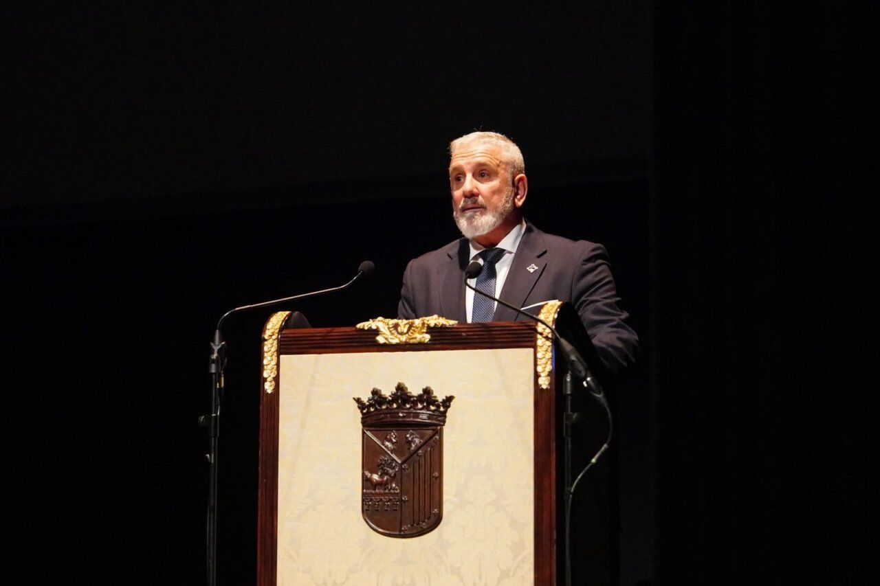 Un hombre habla desde un atril con el escudo de Salamanca sobre un escenario oscuro.