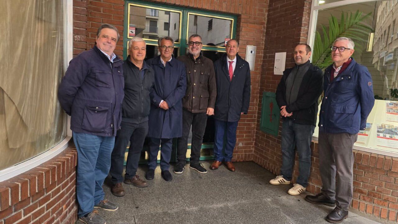 Ocho hombres posan frente a la entrada acristalada de un edificio de ladrillo.