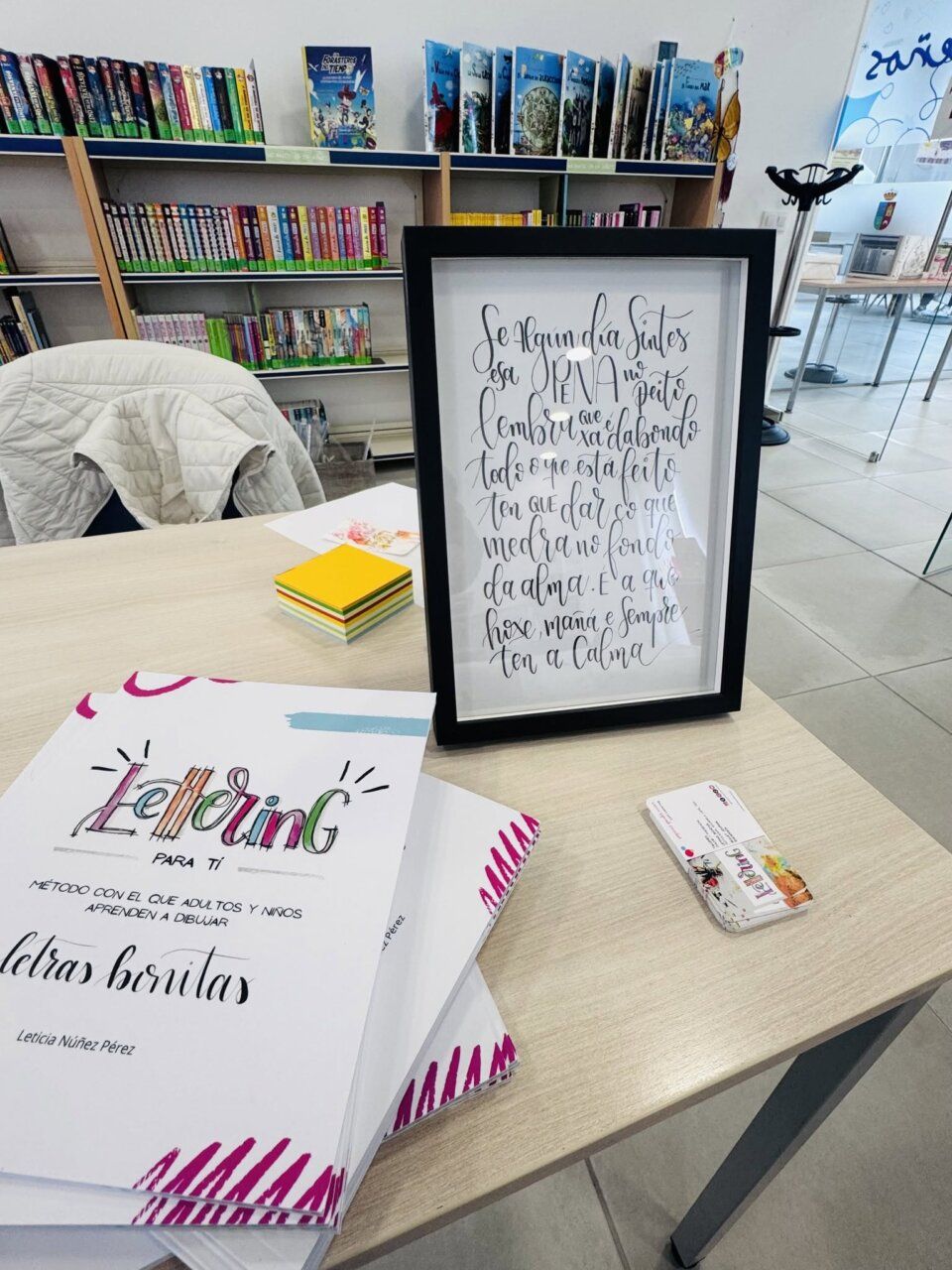 Libros de lettering y láminas caligráficas sobre una mesa frente a estanterías.