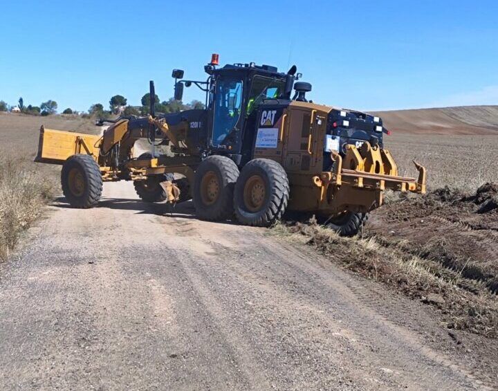 Una motoniveladora amarilla está sobre un camino de tierra entre campos abiertos.