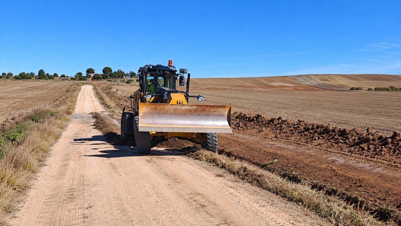Una motoniveladora amarilla trabaja sobre un camino de tierra entre campos.