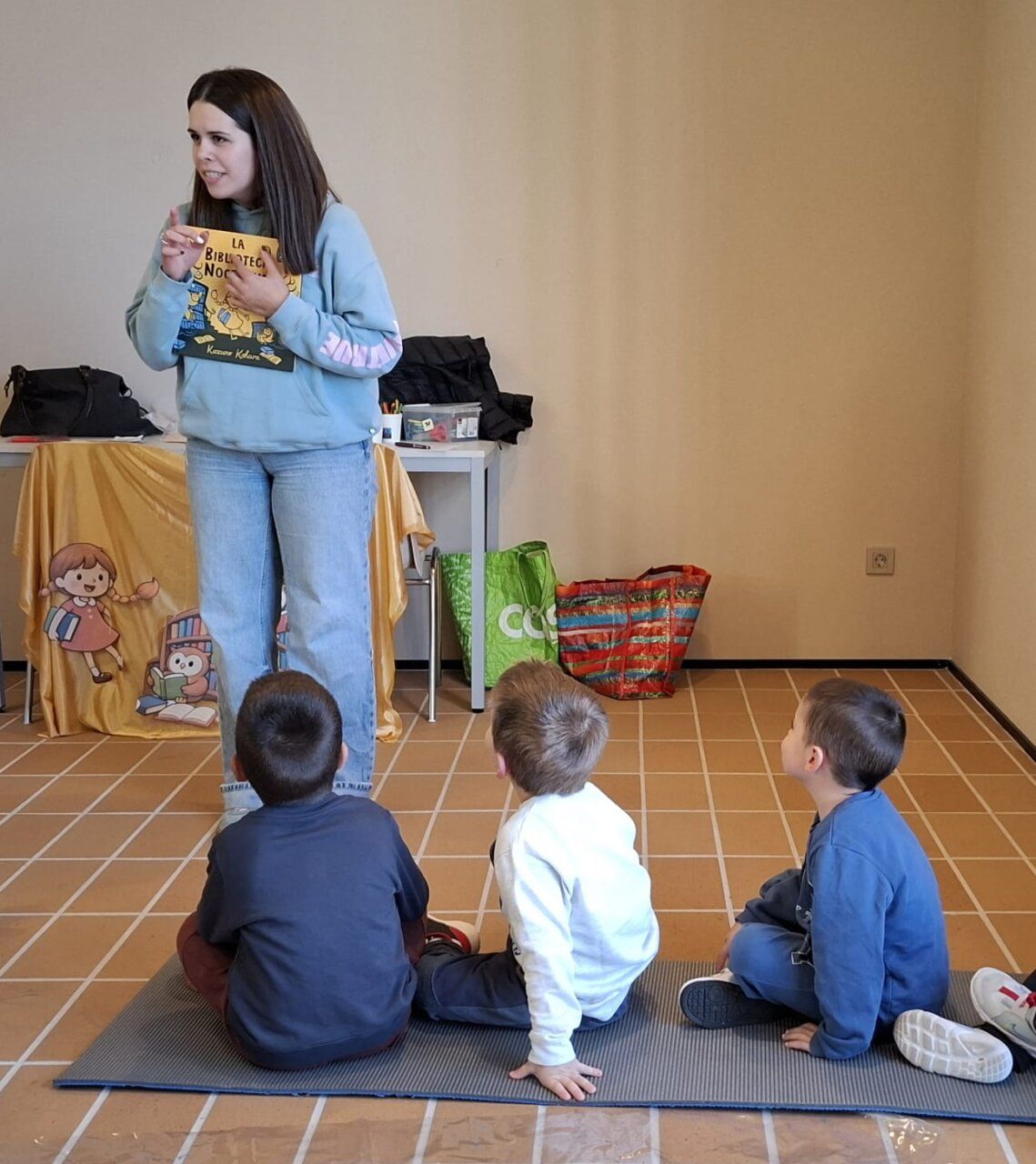 Una mujer sostiene un libro ilustrado frente a tres niños sentados en el suelo.