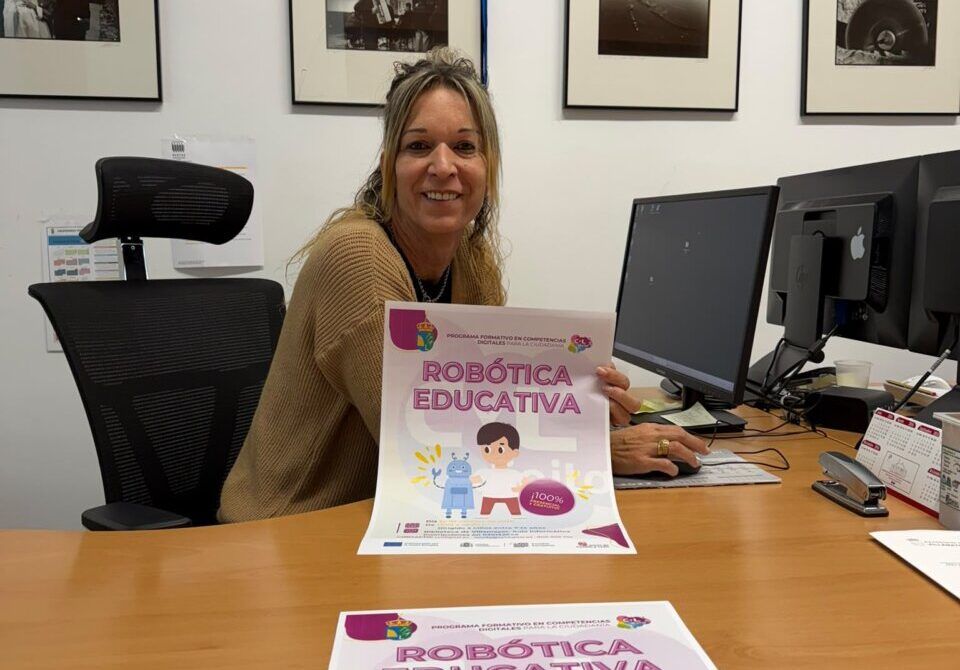 Una mujer muestra un cartel de robótica educativa sentada frente a un escritorio.