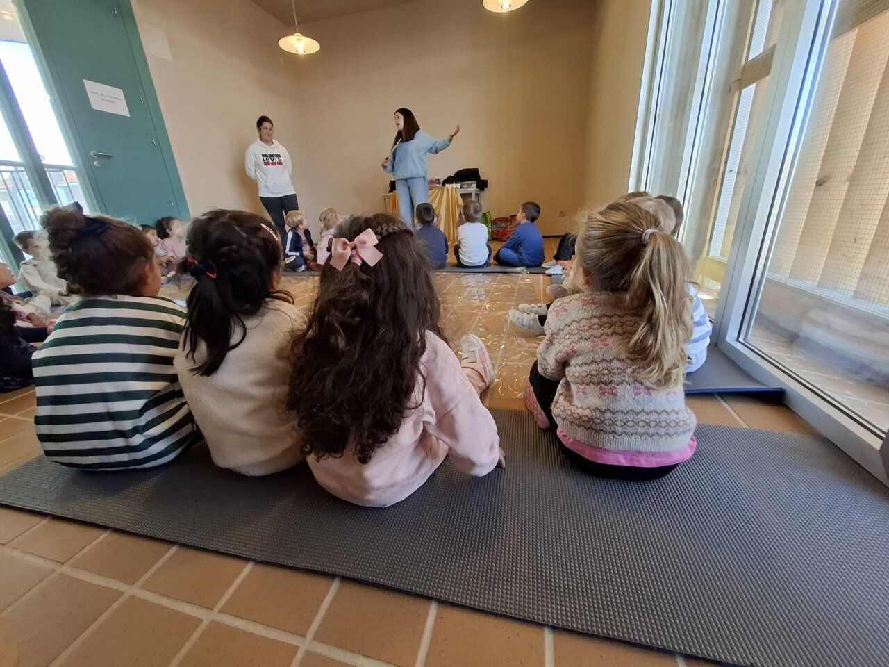 Niños sentados en el suelo escuchan a una mujer que gesticula en una sala.