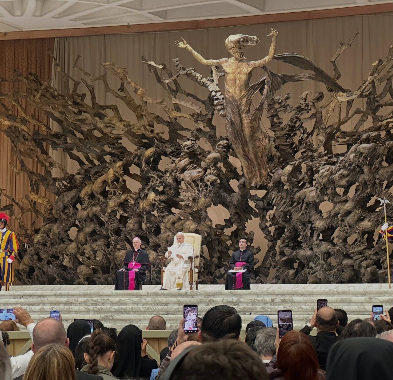 Persona vestida de blanco sentada entre dos clérigos ante una gran escultura y público