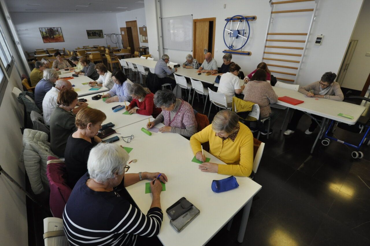 Personas mayores escriben en hojas alrededor de varias mesas en una sala amplia.