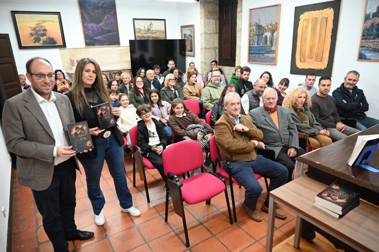 Dos personas muestran un libro ante un grupo sentado en una sala con cuadros.