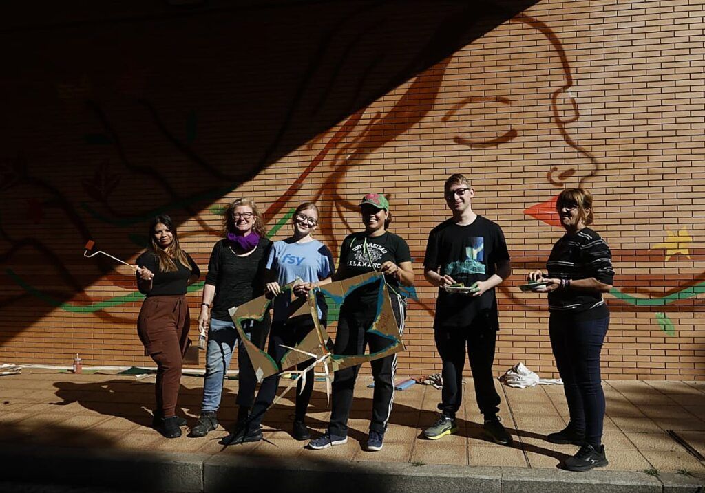 Seis personas posan ante un mural de un rostro femenino pintado sobre ladrillo.