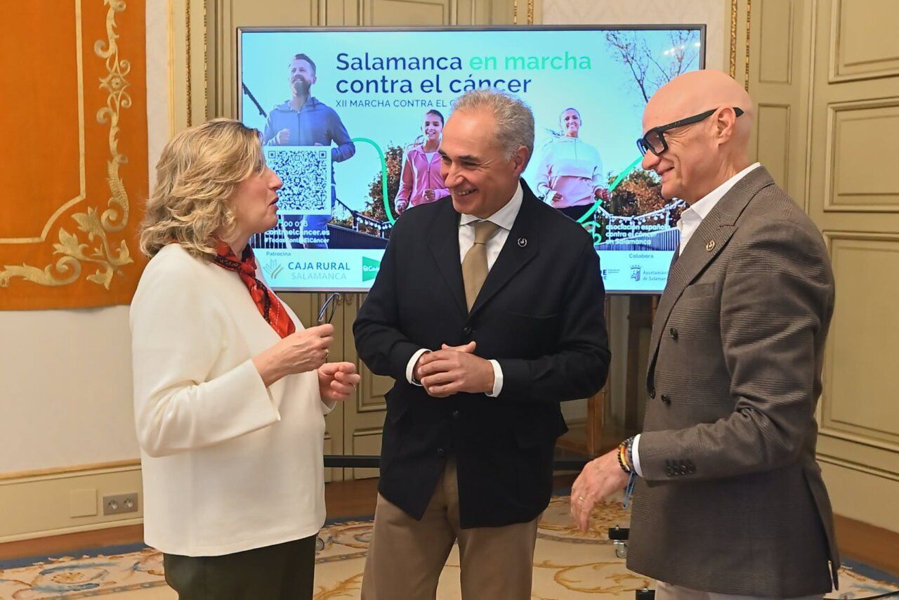 Tres personas conversan ante una pantalla de la XII Marcha contra el Cáncer en Salamanca.