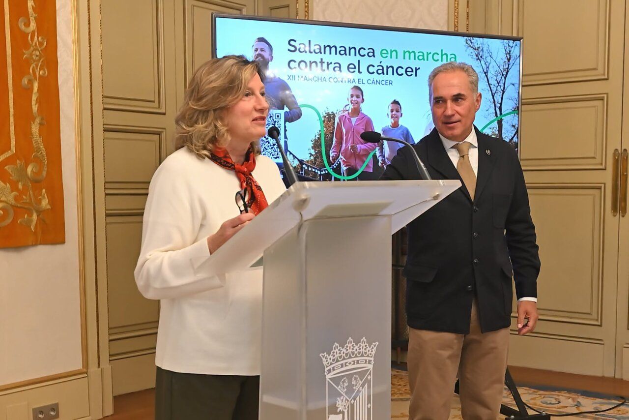Una mujer habla desde un atril junto a un hombre, con cartel de marcha detrás.