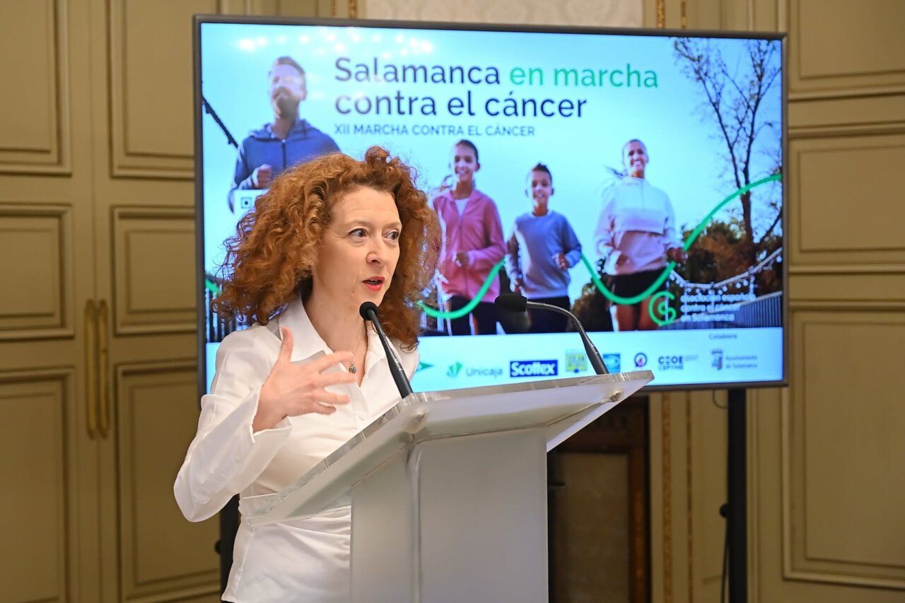 Mujer habla desde un atril ante la pantalla de la XII Marcha contra el Cáncer