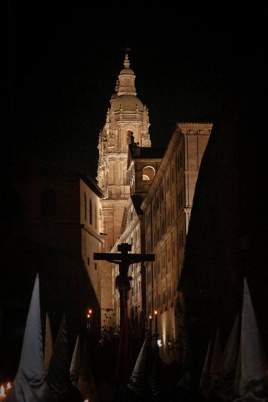 Cruz de procesión y capirotes en una calle nocturna ante una torre iluminada
