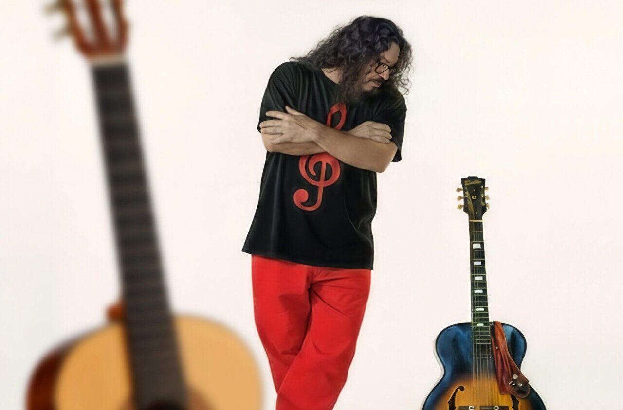 Hombre de cabello rizado, con ropa negra y roja, junto a dos guitarras.