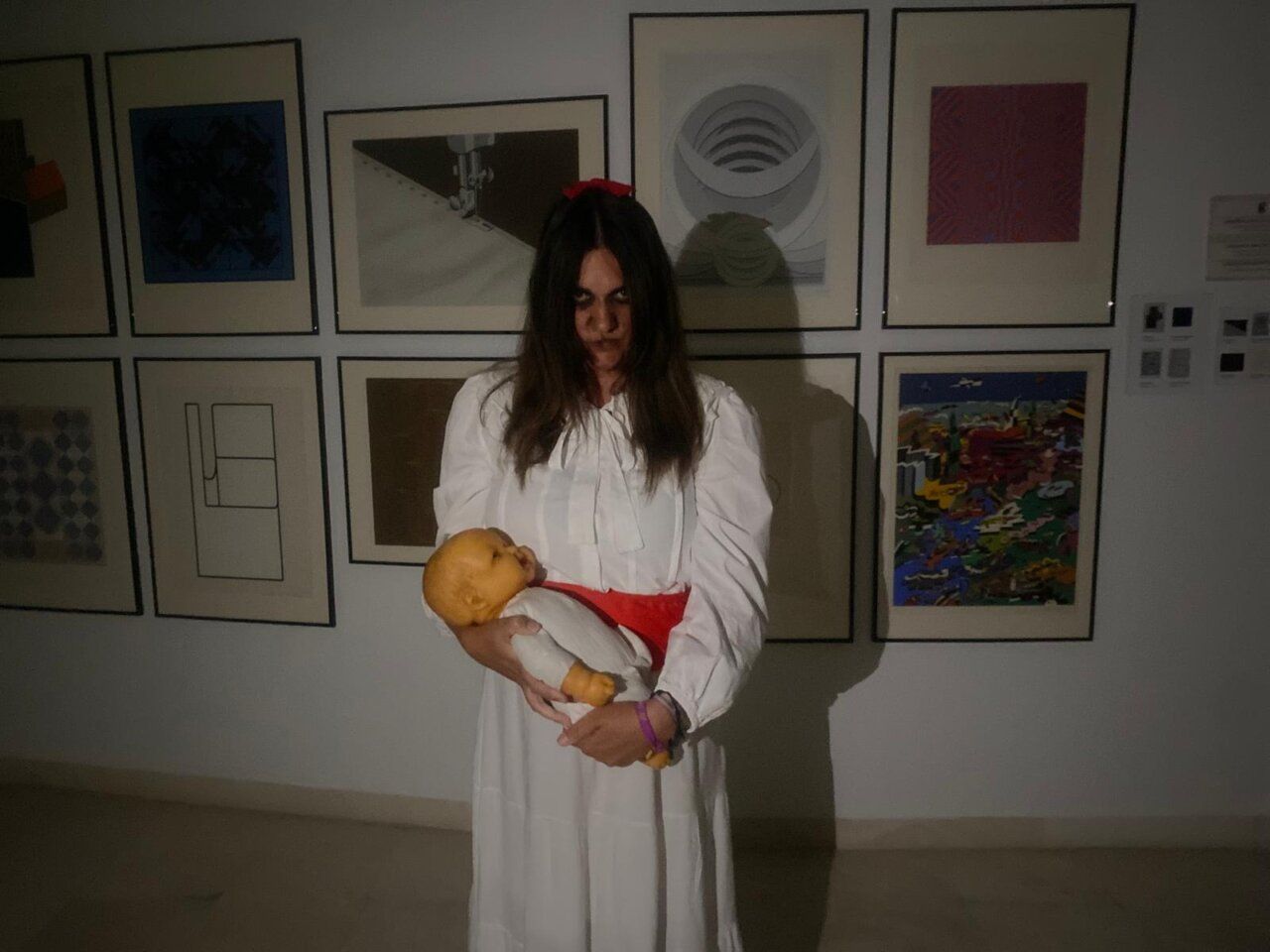 Persona con maquillaje oscuro sostiene un muñeco ante obras enmarcadas en una sala de exposición.