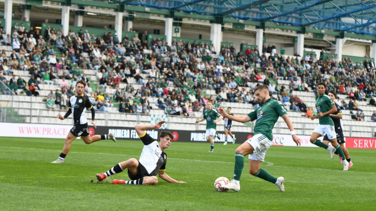 Fotografía de un jugador de verde con el balón ante un rival que cae al césped.
